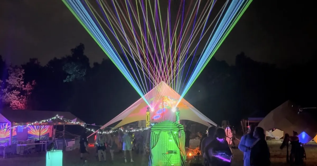 The Cosmic Creatures camp at night at Flipside: rainbow lasers fan out above a lit stage tent while people gather and dance